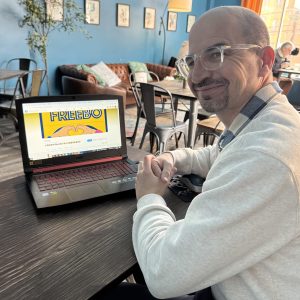 man with glasses and white shirt smiles in coffee shop with laptop