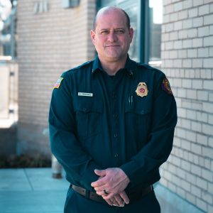 A portrait of a man in a fireman's uniform standing in front of the municipal building