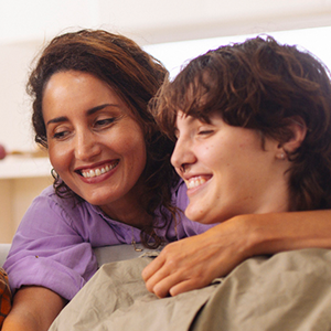 A close up of a mom with dark hair wearing a purple top and her son wearing a beige shirt smiling and laughing together.