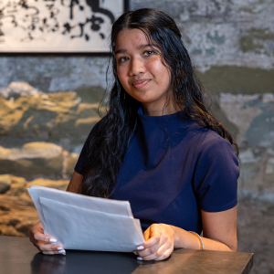 A young woman with long dark hair wearing a short sleeved navy blue dress sits at a table holding papers with both hands in front of a gray wall with an abstract pattern and the corner of a black and white piece of art.