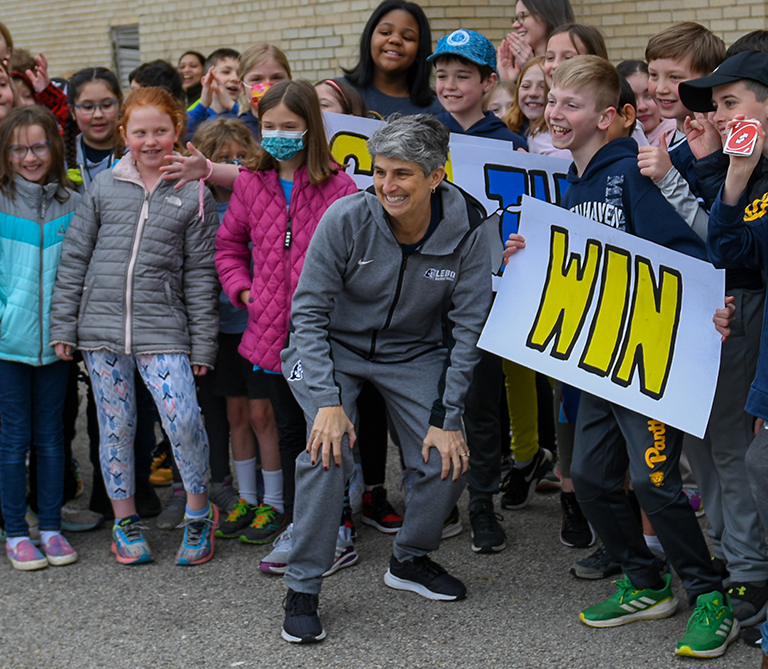 An excited group of children holding white signs with yellow and blue block letters outlined in black that spell "WIN" stand behind a coach in a grey tracksuit.