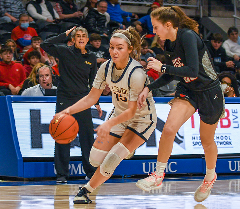 Basketball game action shot showing a player in a white uniform dribbling while being closely guarded by an opponent in a navy blue uniform. A coach is standing in the background with her right arm raised above her head in front of the announcers box and a large crowd seated in the stands.