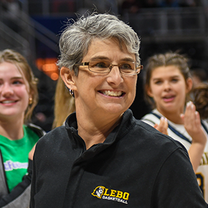 Team coach with short gray hair and glasses smiling and wearing a black jacket with a gold and white "Lebo Basketball" team logo, members of the team can be seen in the background smiling and clapping.