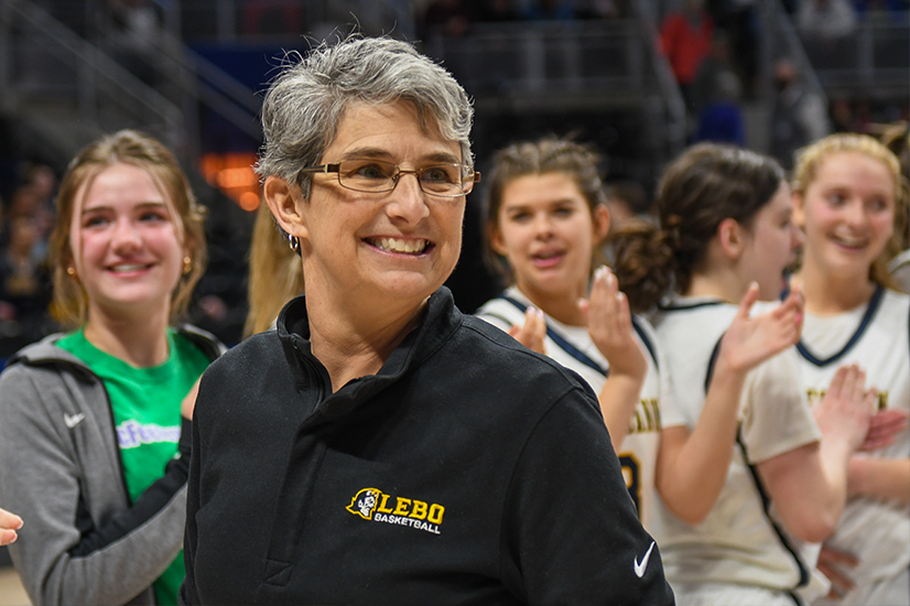 Team coach with short gray hair and glasses smiling and wearing a black jacket with a gold and white "Lebo Basketball" team logo, members of the team can be seen in the background smiling and clapping.