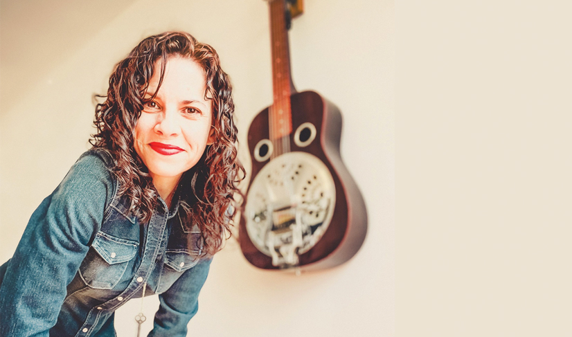 A woman wearing a jean jacket, standing, leaning over looking at the camera and there is a steel guitar hangin on the wall behind her