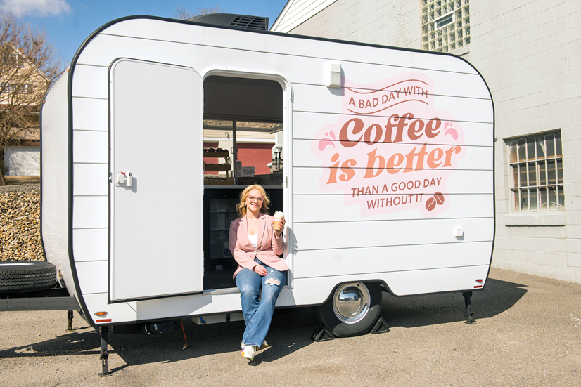 A blonde haired woman sitting in a trailer with a coffee cop, there is a quote on the trailer that says "a bad day with coffee is better than a good day without it."