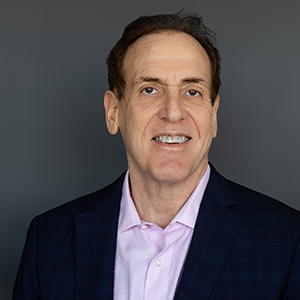 A man with short dark brown hair smiles while wearing a dark blazer and light pink shirt in front of a gray background.