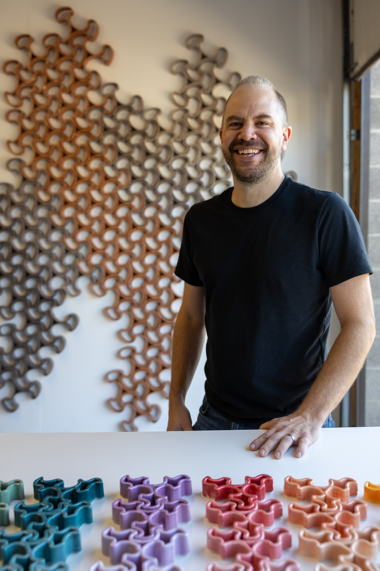 Man in a black shirt smiling, standing behind a table with colorful geometric shapes with Intricate wall art in brown tones in the background.