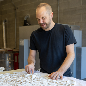 A person wearing a black t-shirt looking down while working on an intricate white 3D model on a table. Gray cinder blocks and industrial looking equipment can be seen in the background.