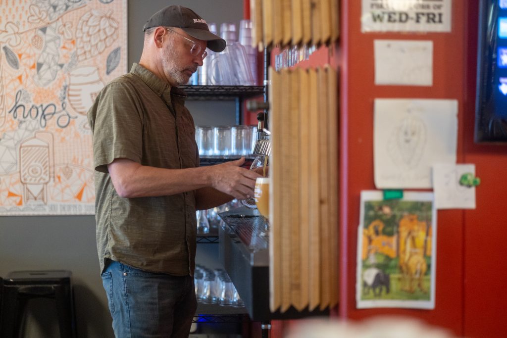 man with green short sleeve shirt and hat pouring beer from a tap in a bar