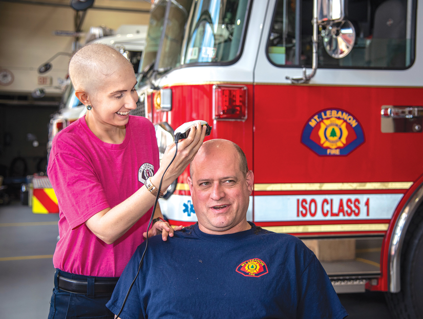 A cancer patient woman with shaved head shaving a mans head next to fire trucks