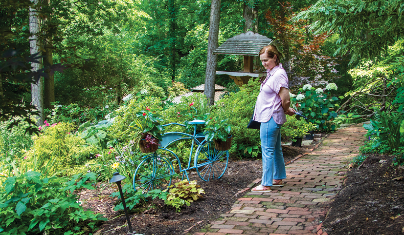 A woman standing on a brick path in a garden surrounded by greenery next to a bike on display looking at a bush