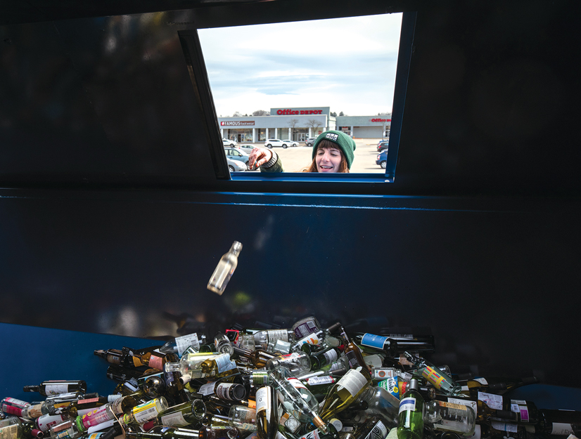 a view from inside a recycling bin of a woman throwing a bottle into the bin with a pile of glass bottles