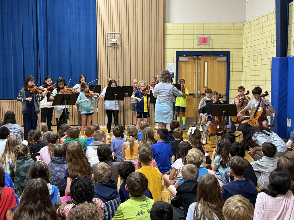 children in a school gymnasium sitting in a crowd, while other kids and a teacher perform with instruments