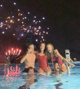 five teenage lifeguards pose in pool at dark with fireworks in background