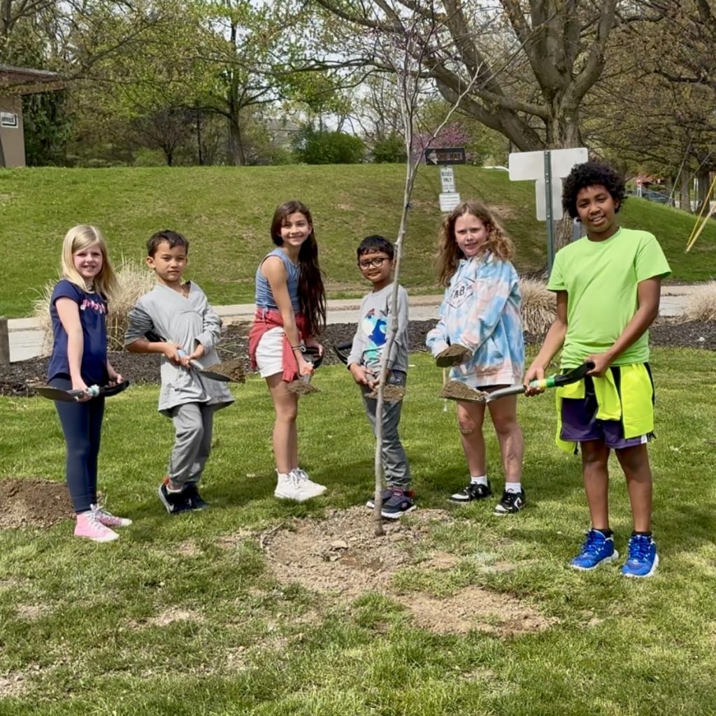 six elementary school kids stand with shovels in front of a newly planted tree