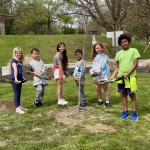 six elementary school kids stand with shovels in front of a newly planted tree