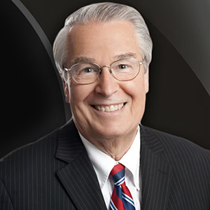 Portrait of an older man with gray hair and glasses smiling and wearing a dark pinstripe suit, white button down shirt and red and blue striped tie.