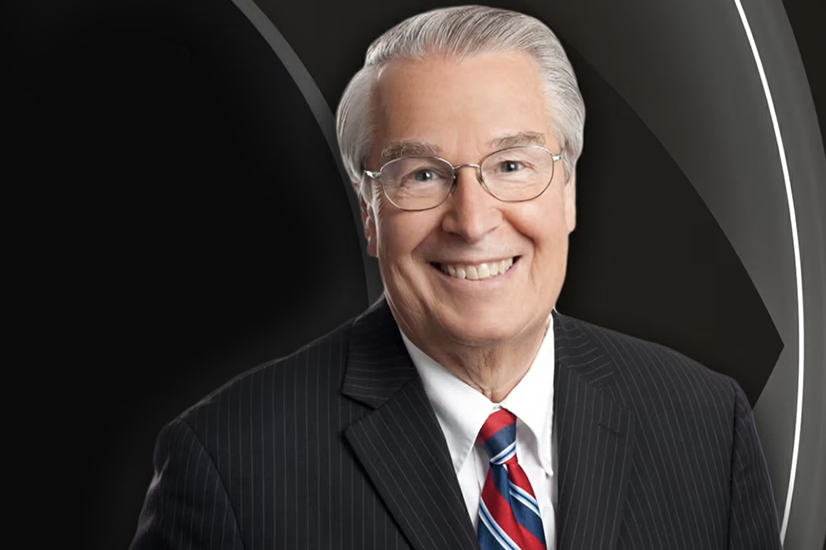 Portrait of an older man with gray hair and glasses smiling and wearing a dark pinstripe suit, white button down shirt and red and blue striped tie.