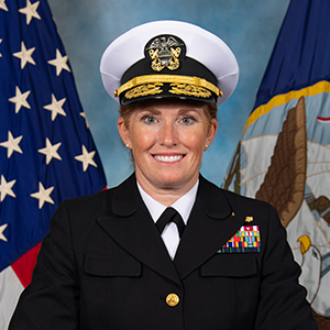 A woman in U.S. Navy uniform stands smiling against a backdrop of the American and Navy flags.