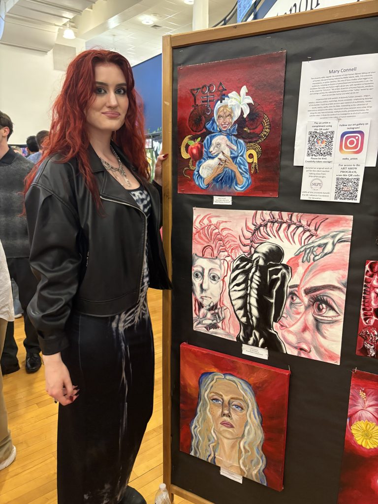 young woman with long red hair and all black clothes stands by her artwork display in an art show