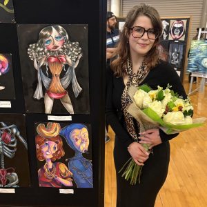 young woman with glasses and long brain hair in black clothes holding flowers smiles by artwork display with painted woman with an apple for a torso