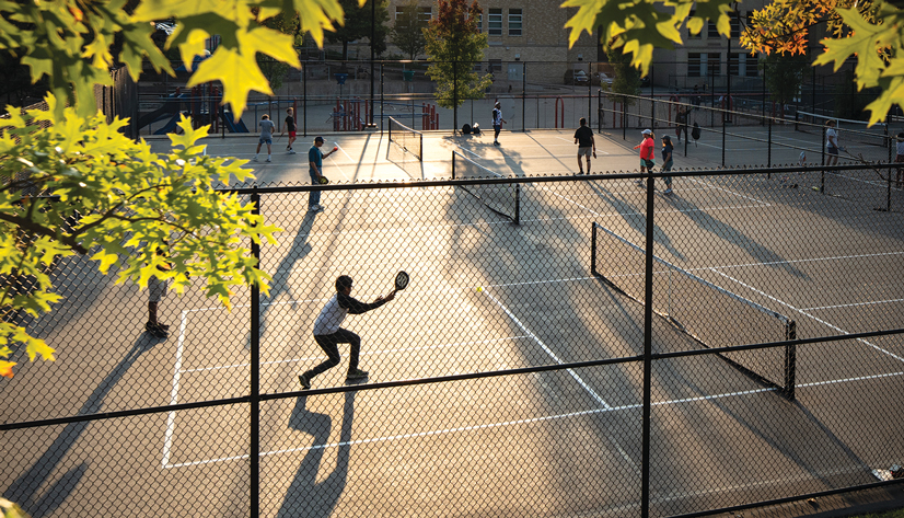 Several people playing pickleball on the pickleball courts, the sum is shining through the trees and there is long shadows across the court