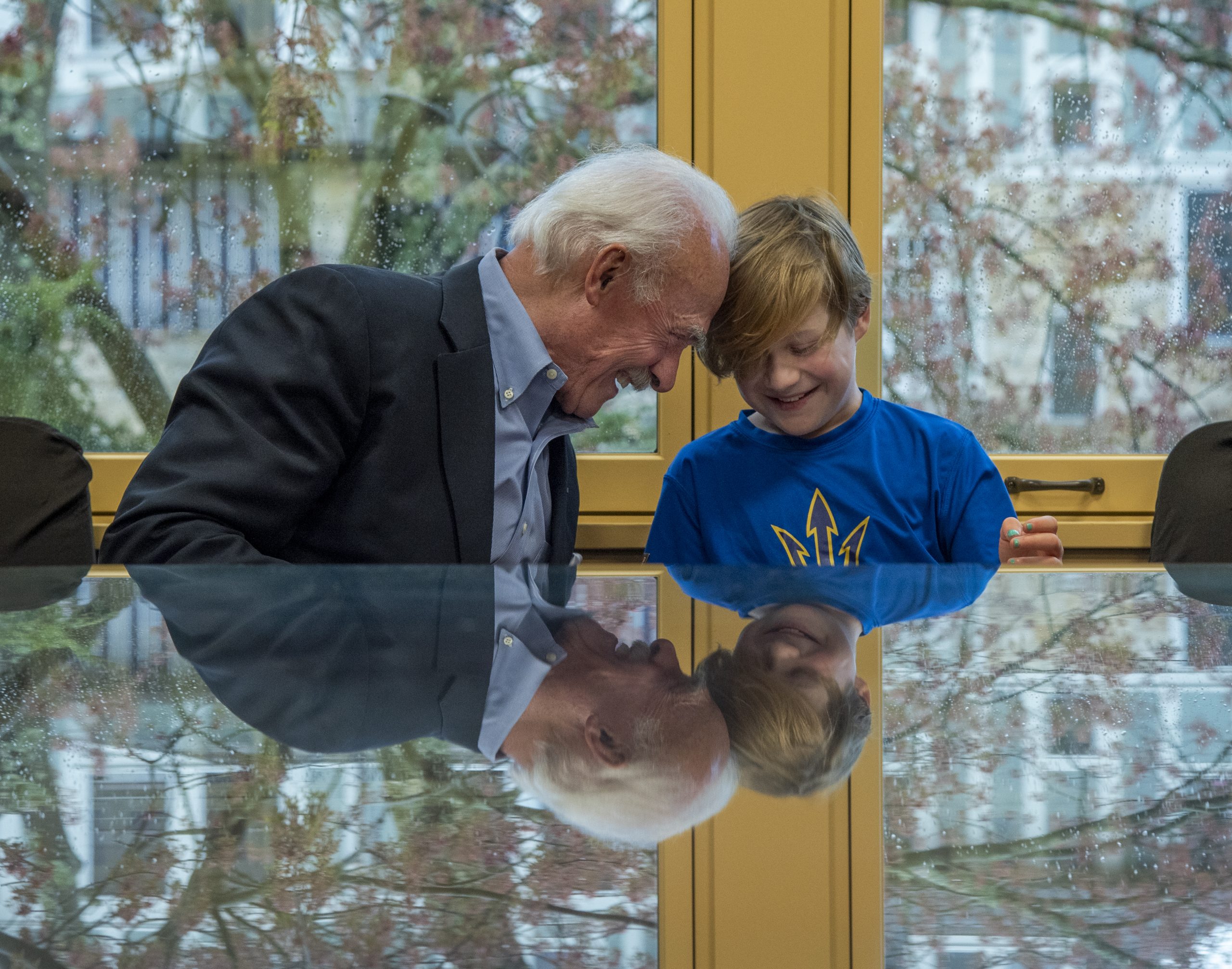 elderly man with suit and white hair sitting by a young boy with a blue shirt and blonde hair, both smiling