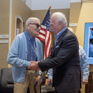 two elderly men shake hands in front of American flag