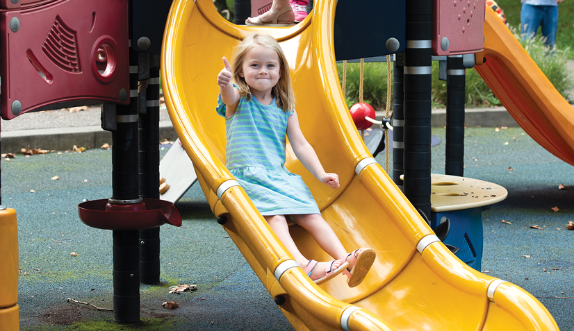 A young girl wearing a blue dress sliding down a slide on a playground, smiling, giving the thumbs up 