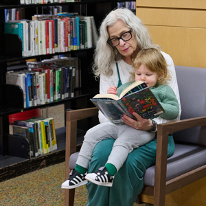A woman with grey hair and glasses reading to a child sitting on her lap looking at a book