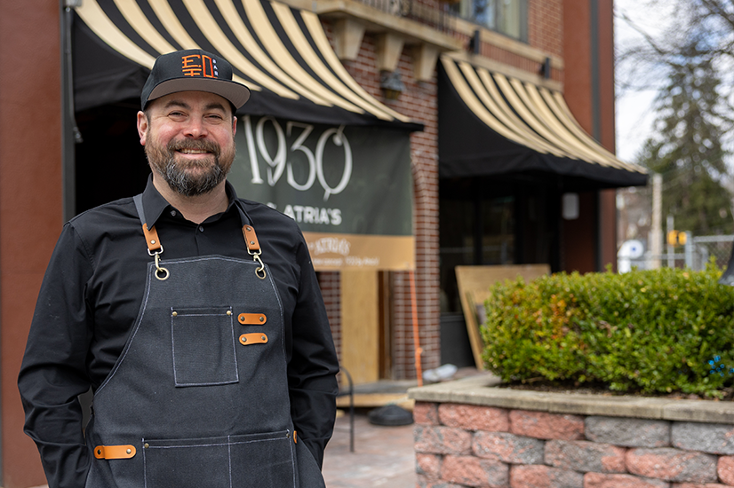 Man in a black button down shirt and black baseball hat with an orange design, wearing a dark apron with leather straps, smiles while standing in front of a red brick building with gold and black striped awnings over the windows and a large black sign with "1930 by Atria’s” in white text.