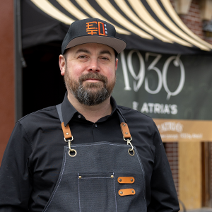 Man in a black button down shirt and black baseball hat with an orange design, wearing a dark apron with leather straps, stands in front of a red brick building with gold and black striped awnings over the windows and a large black sign with "1930 by Atria’s” in white text.
