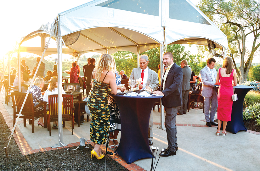 people in dress attire standing outside under a tent, drinking and eating food at a fundraiser