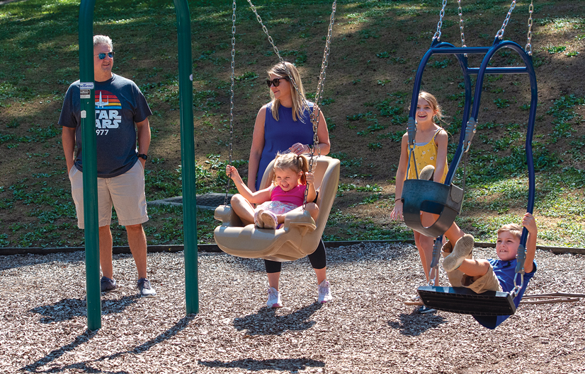 a family, a man and woman and three kids, at a park, and two of the kids are swinging on swings.