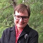 woman with short brown hair, glasses and black jacket smiles in front of green tree