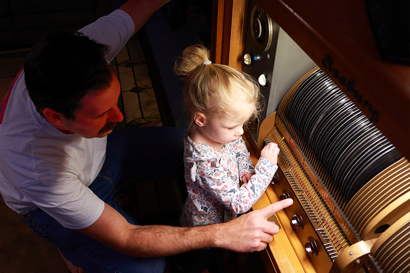 A man with short dark hair and a mustache wearing a white t-shirt crouches behind a young girl with blonde hair in a pony tail and a long sleeved shirt with small flower pattern as they look at a vintage jukebox with a line of records behind the glass front.