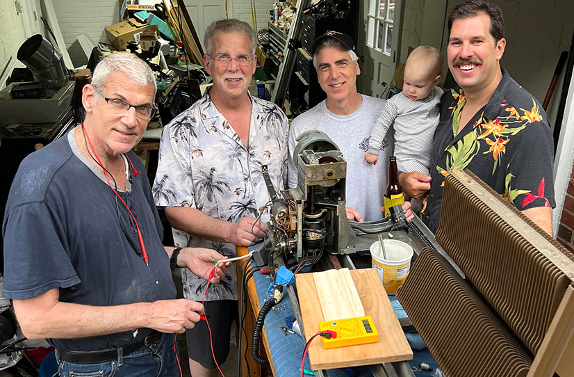 Four men standing together (the last one on the right is holding a baby) smiling in a garage behind a variety of vintage equipment and pieces from a jukebox.