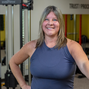 A woman with shoulder length dark blonde hair wearing a sleeveless gray top stands in a gym smiling with one hand on her hip and the other on a treadmill. A variety of exercise equipment is visible in the background.