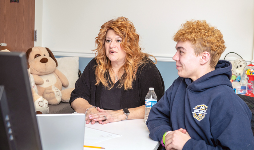 woman with curly red/blonde hair and black shirt talking at table with teen wearing hoodie