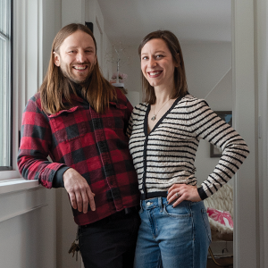 A couple smiling and standing together in a hallway. The man has long light brown hair and is wearing a red plaid button down shirt. The woman has brow shoulder length hair and is wearing a striped black and white sweater and jeans.
