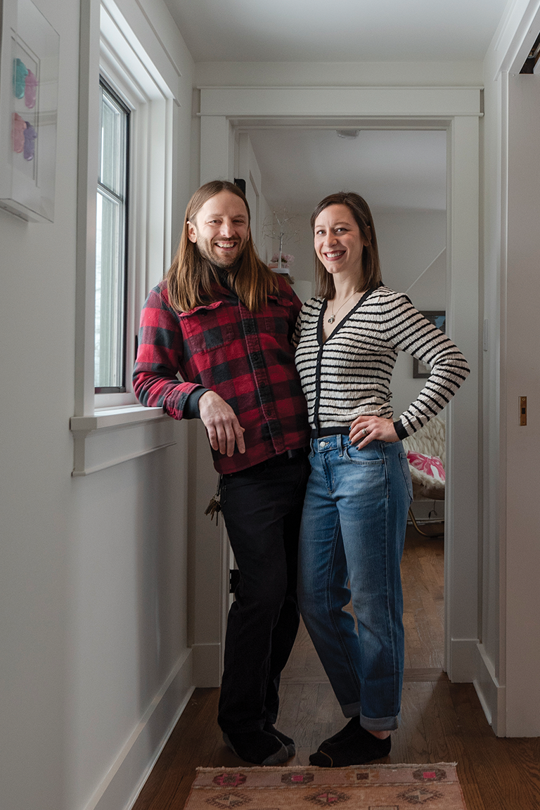 A couple smiling and standing together in a hallway. The man has long light brown hair and is wearing a red plaid button down shirt. The woman has brow shoulder length hair and is wearing a striped black and white sweater and jeans.