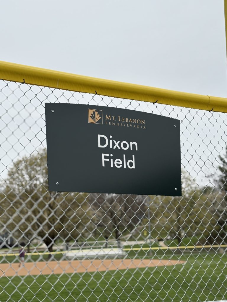 green and white sign that reads dixon field on a fence at a baseball field