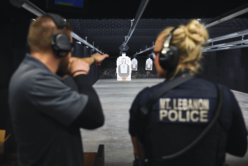 A man standing next to a woman police officer at a firing range, pretending to hold a rifle while she looks at him
