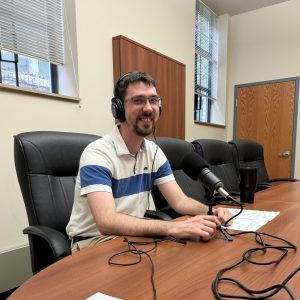 Ian McMeans sits at desk in front of microphone, wearing headphones.