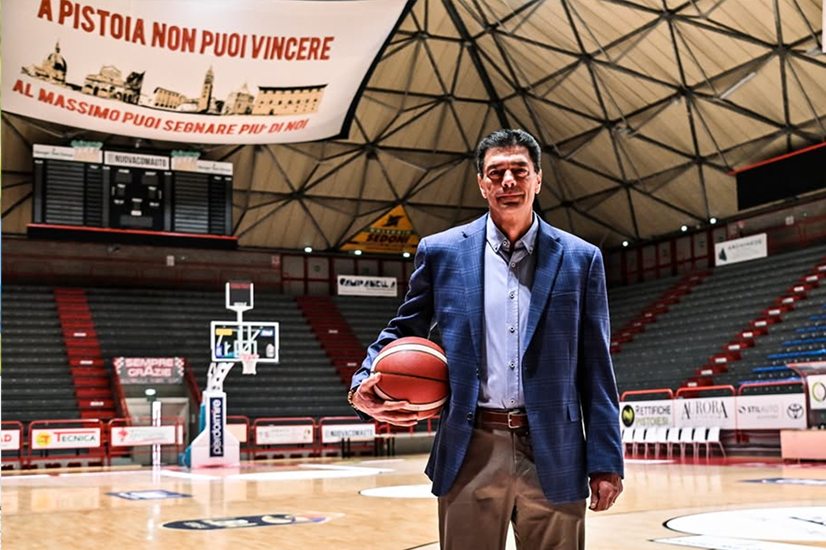 A man in a blue blazer holds a basketball while standing on an empty indoor court. The arena seats, a large banner and a basketball hoop can be seen in the background.