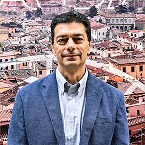 A headshot style image of a man with short dark hair in a blue blazer on top of a wide shot of a city scape in Italy.