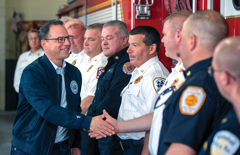 man with glasses shaking hands of emergency service workers with a fire truck in the background