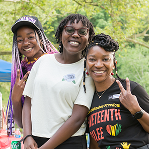 Three black people standing together and smiling in a park. The person on the left is wearing a black baseball hat and has long braids dyed with a purple ombre. The person in the middle has short hair and is wearing glasses and a white t-shirt. The person on the right is showing a peace sign and is wearing a black t-shirt with the words Celebrates Freedom (in yellow letters) and Juneteenth (in red letters.)