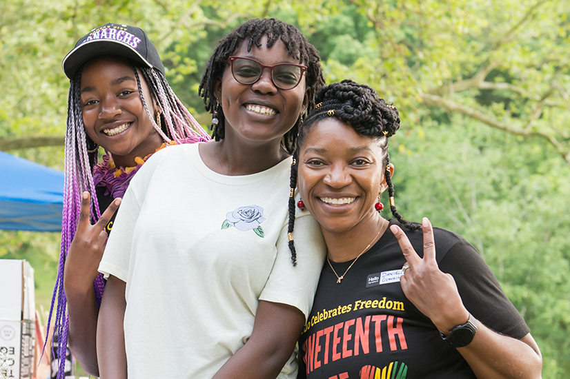 Three black people standing together and smiling in a park. The person on the left is wearing a black baseball hat and has long braids dyed with a purple ombre. The person in the middle has short hair and is wearing glasses and a white t-shirt. The person on the right is showing a peace sign and is wearing a black t-shirt with the words Celebrates Freedom (in yellow letters) and Juneteenth (in red letters.)